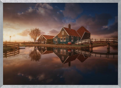 A picturesque landscape of traditional Dutch houses with teal walls and red roofs reflected perfectly in the calm water of a canal at sunset, under a dramatic, cloudy sky. A small wooden bridge crosses the canal, and the scene is enclosed in a silver frame. Artwork