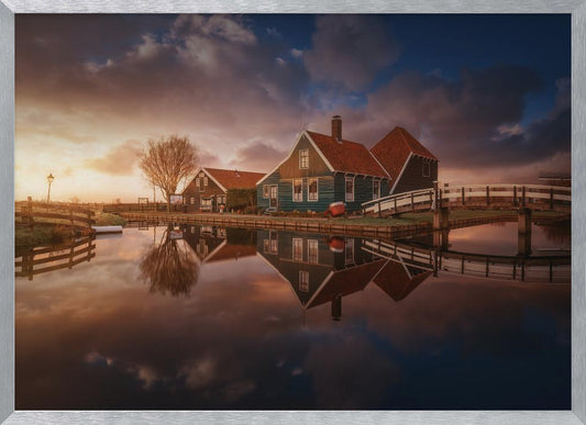 A picturesque landscape of traditional Dutch houses with teal walls and red roofs reflected perfectly in the calm water of a canal at sunset, under a dramatic, cloudy sky. A small wooden bridge crosses the canal, and the scene is enclosed in a silver frame. Artwork