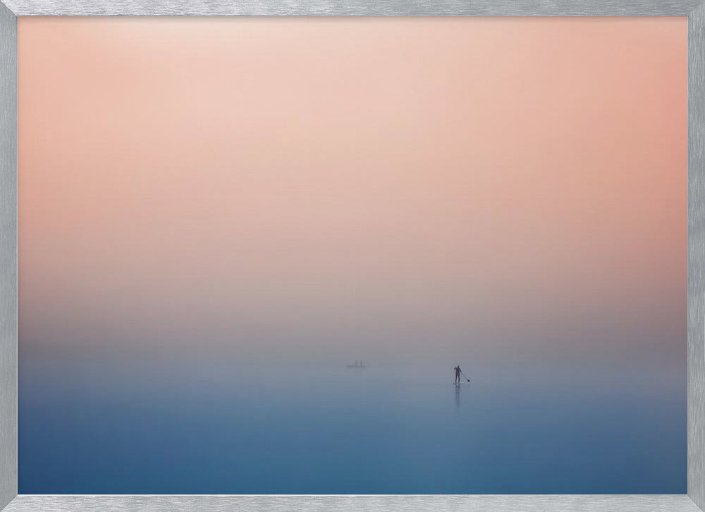 A minimalist framed photograph of a solitary person on a stand-up paddleboard, silhouetted against a vast, calm expanse of water under a hazy, gradient sky that shifts from soft pink to deep blue. Wall Art