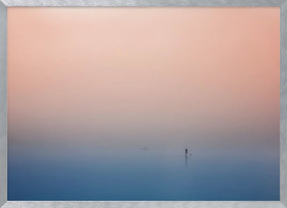 A minimalist framed photograph of a solitary person on a stand-up paddleboard, silhouetted against a vast, calm expanse of water under a hazy, gradient sky that shifts from soft pink to deep blue. Wall Art