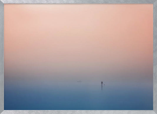 A minimalist framed photograph of a solitary person on a stand-up paddleboard, silhouetted against a vast, calm expanse of water under a hazy, gradient sky that shifts from soft pink to deep blue. Wall Art