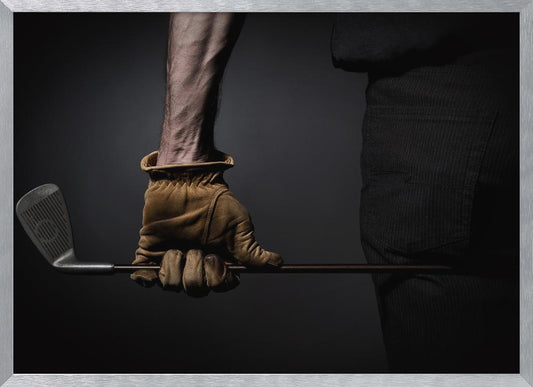 A dramatic close-up of a person's muscular forearm and hand wearing a tan leather glove, holding a golf iron horizontally against a dark background, framed in silver. Wall Art