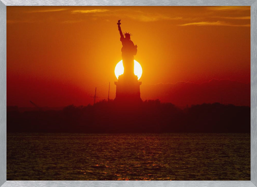 A powerful silhouette of the Statue of Liberty against a fiery orange and red sunset. The sun is perfectly positioned behind the statue's base, creating a brilliant, glowing orb of light. The dark waters in the foreground reflect the vibrant colors of the sky. The photograph is presented within a silver-colored frame. Artwork