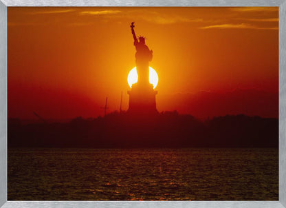 A powerful silhouette of the Statue of Liberty against a fiery orange and red sunset. The sun is perfectly positioned behind the statue's base, creating a brilliant, glowing orb of light. The dark waters in the foreground reflect the vibrant colors of the sky. The photograph is presented within a silver-colored frame. Artwork