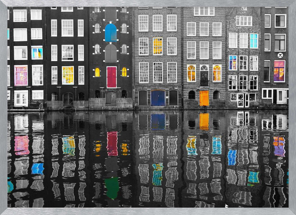 A framed black and white photograph of canal houses in Amsterdam, with selective coloring highlighting windows and doors in vibrant shades of red, blue, yellow, and pink. The colorful architectural details are mirrored in the rippling water of the canal below. Artwork