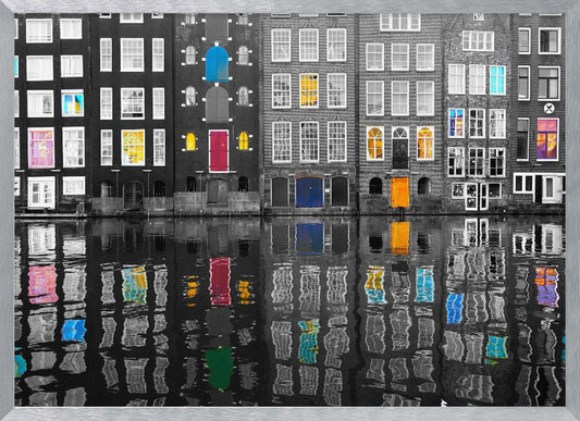 A framed black and white photograph of canal houses in Amsterdam, with selective coloring highlighting windows and doors in vibrant shades of red, blue, yellow, and pink. The colorful architectural details are mirrored in the rippling water of the canal below. Artwork