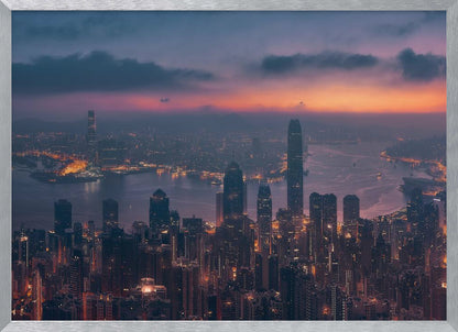 An aerial photograph of a sprawling city skyline at dusk, likely Hong Kong, with countless illuminated skyscrapers. The sky above transitions from a warm orange glow at the horizon to a deep purple, with dark clouds overhead. The city lights cast a golden reflection on the water of the harbor. The entire image is presented within a silver-colored frame. Poster