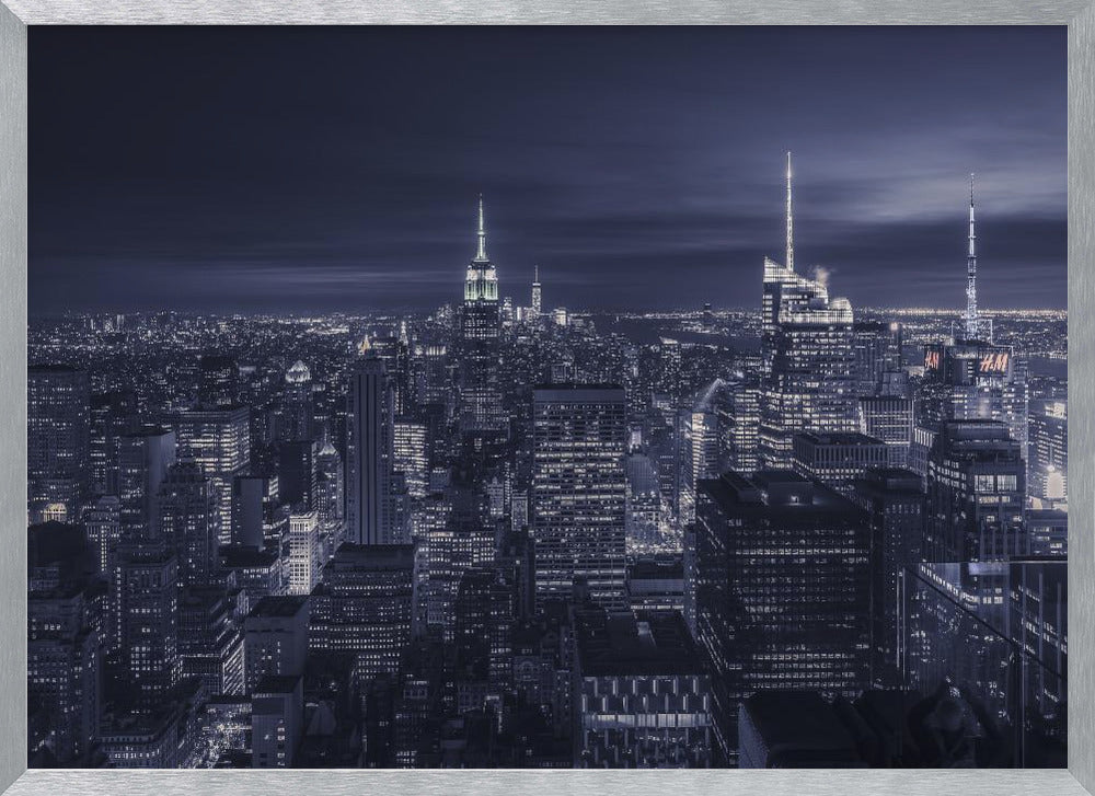 A dramatic nighttime photograph of the New York City skyline, viewed from a high vantage point. The cityscape is a sea of illuminated skyscrapers under a dark, moody sky, with the entire image cast in a cool blue and white monochrome tone and presented in a silver frame. Artwork