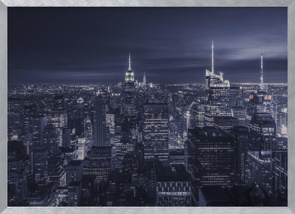 A dramatic nighttime photograph of the New York City skyline, viewed from a high vantage point. The cityscape is a sea of illuminated skyscrapers under a dark, moody sky, with the entire image cast in a cool blue and white monochrome tone and presented in a silver frame. Artwork