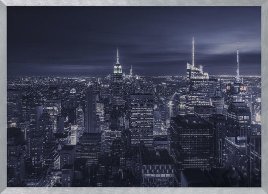 A dramatic nighttime photograph of the New York City skyline, viewed from a high vantage point. The cityscape is a sea of illuminated skyscrapers under a dark, moody sky, with the entire image cast in a cool blue and white monochrome tone and presented in a silver frame. Artwork