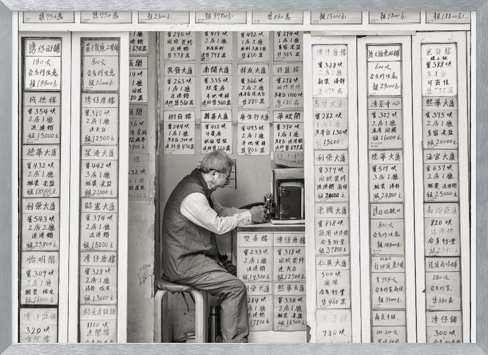 A black and white photograph of an elderly man with glasses sitting on a stool in a small, cramped shop. He is hunched over a counter, operating a small machine. The walls around him are completely covered with vertical paper signs featuring handwritten Chinese calligraphy, likely property listings or advertisements. Artwork