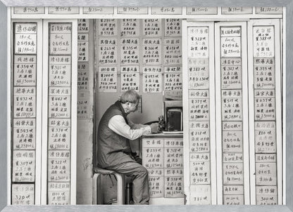 A black and white photograph of an elderly man with glasses sitting on a stool in a small, cramped shop. He is hunched over a counter, operating a small machine. The walls around him are completely covered with vertical paper signs featuring handwritten Chinese calligraphy, likely property listings or advertisements. Artwork