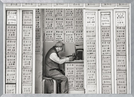 A black and white photograph of an elderly man with glasses sitting on a stool in a small, cramped shop. He is hunched over a counter, operating a small machine. The walls around him are completely covered with vertical paper signs featuring handwritten Chinese calligraphy, likely property listings or advertisements. Artwork