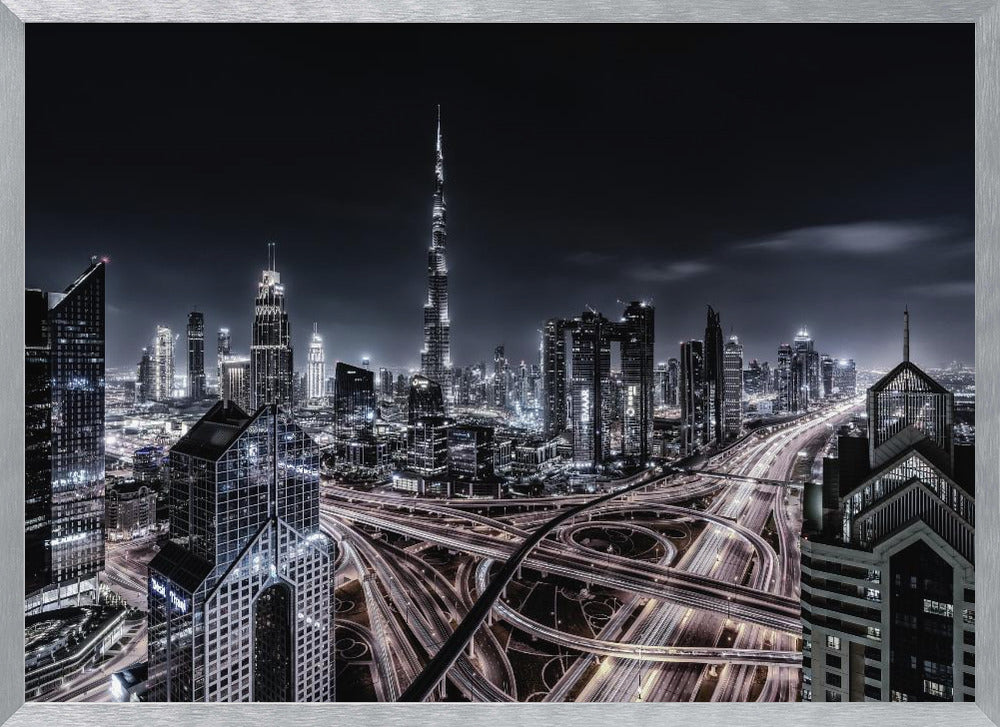 A striking black and white aerial photograph of the Dubai skyline at night, captured in a silver frame. The Burj Khalifa stands tall in the background, while a complex web of illuminated highway overpasses and light trails from traffic fills the foreground, creating a dynamic and futuristic cityscape. Print