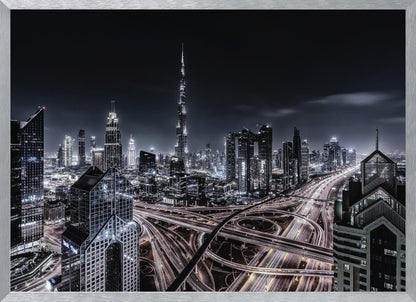 A striking black and white aerial photograph of the Dubai skyline at night, captured in a silver frame. The Burj Khalifa stands tall in the background, while a complex web of illuminated highway overpasses and light trails from traffic fills the foreground, creating a dynamic and futuristic cityscape. Print