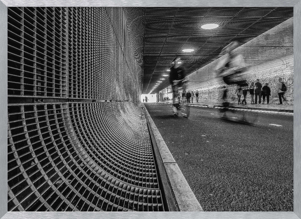 A dynamic black and white photograph from inside a modern tunnel. On the left, a curved metal grate wall creates a strong pattern, while on the right, cyclists are captured in motion blur on a paved path. Pedestrians are visible in the background, walking through the illuminated passage. Poster