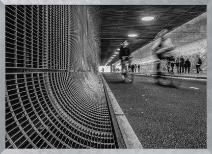 A dynamic black and white photograph from inside a modern tunnel. On the left, a curved metal grate wall creates a strong pattern, while on the right, cyclists are captured in motion blur on a paved path. Pedestrians are visible in the background, walking through the illuminated passage. Poster