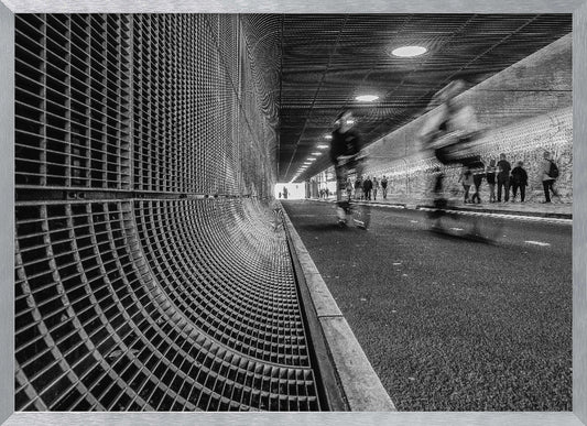A dynamic black and white photograph from inside a modern tunnel. On the left, a curved metal grate wall creates a strong pattern, while on the right, cyclists are captured in motion blur on a paved path. Pedestrians are visible in the background, walking through the illuminated passage. Poster