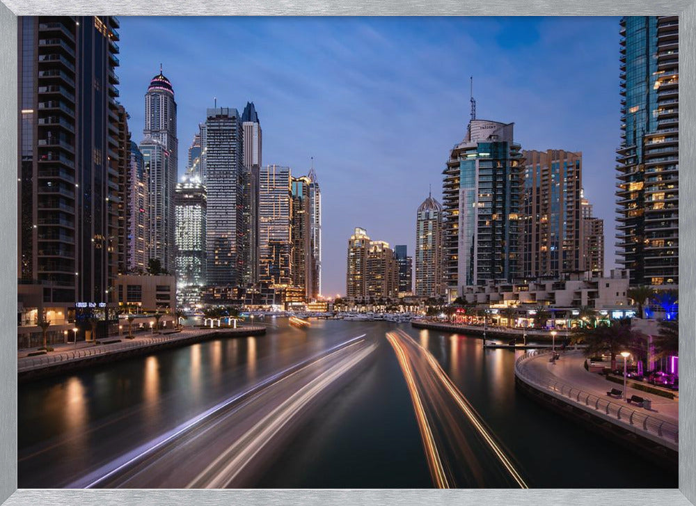 A long-exposure photograph of the Dubai Marina at dusk, framed in silver. Illuminated skyscrapers line a busy waterway, with light trails from moving boats creating long, bright streaks on the water. The sky is a deep twilight blue, and the city's lights cast a warm glow. Decor