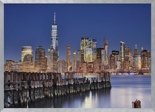 A framed photograph of the Lower Manhattan skyline at dusk. The One World Trade Center and other skyscrapers are brightly lit against a deep blue sky. In the foreground, an old wooden pier extends into the water, which reflects the city lights. Wall Art