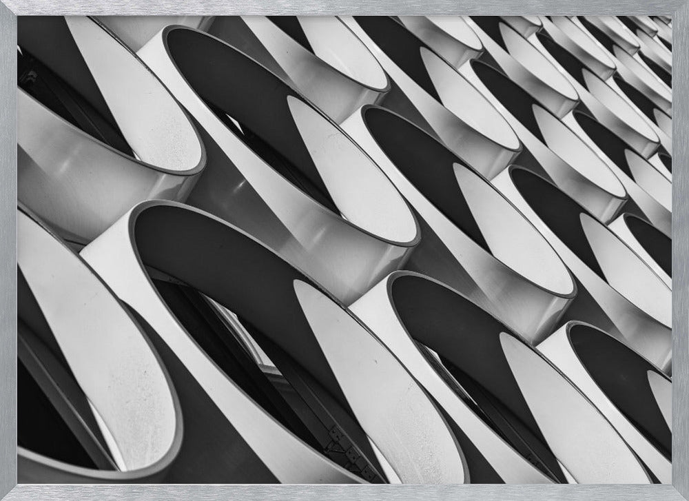 A framed, black and white photograph captures a low-angle, close-up view of a modern building's facade. The image features a repeating pattern of slanted, oval, or teardrop-shaped metallic elements, creating a dynamic interplay of light, shadow, and reflection. Print