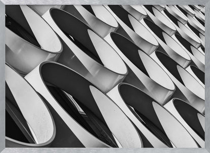 A framed, black and white photograph captures a low-angle, close-up view of a modern building's facade. The image features a repeating pattern of slanted, oval, or teardrop-shaped metallic elements, creating a dynamic interplay of light, shadow, and reflection. Print