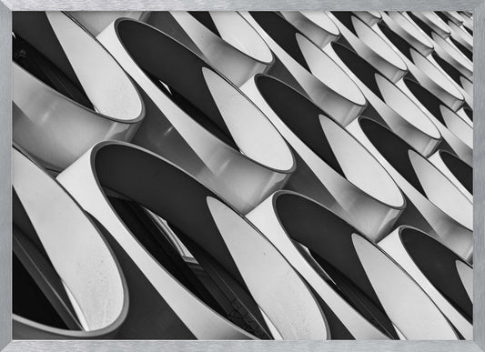 A framed, black and white photograph captures a low-angle, close-up view of a modern building's facade. The image features a repeating pattern of slanted, oval, or teardrop-shaped metallic elements, creating a dynamic interplay of light, shadow, and reflection. Print