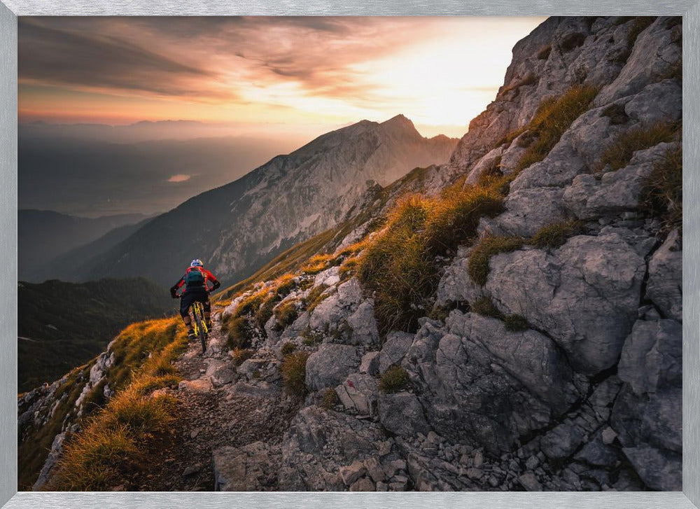 A mountain biker from behind rides along a narrow, rocky trail on a steep mountain ridge during a dramatic golden sunset. In the background, layers of mountains fade into the distance under a warm, glowing sky. Artwork