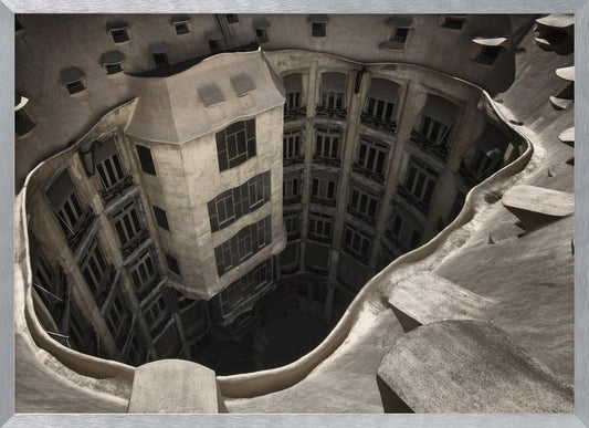 A surreal, high-angle photograph looking down into the distorted, cavernous courtyard of a building. The walls and windows appear to melt and curve inwards towards a dark void, creating a powerful optical illusion. The image has a muted, sepia-toned color palette and is presented in a silver frame. Wall Art