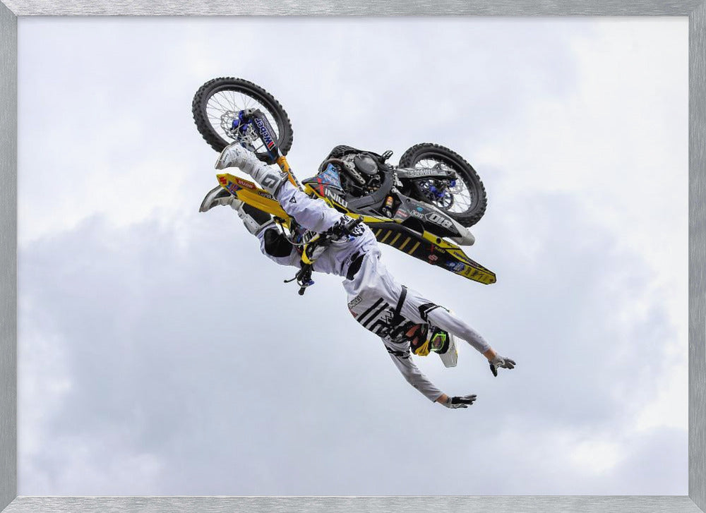 A framed action shot of a freestyle motocross rider in mid-air, performing an upside-down trick against a cloudy sky, with their body hanging below the yellow and black dirt bike. Artwork