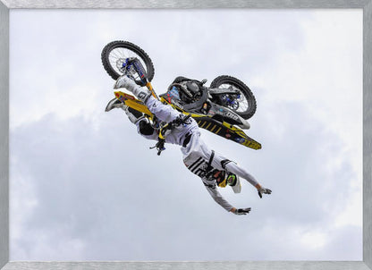 A framed action shot of a freestyle motocross rider in mid-air, performing an upside-down trick against a cloudy sky, with their body hanging below the yellow and black dirt bike. Artwork