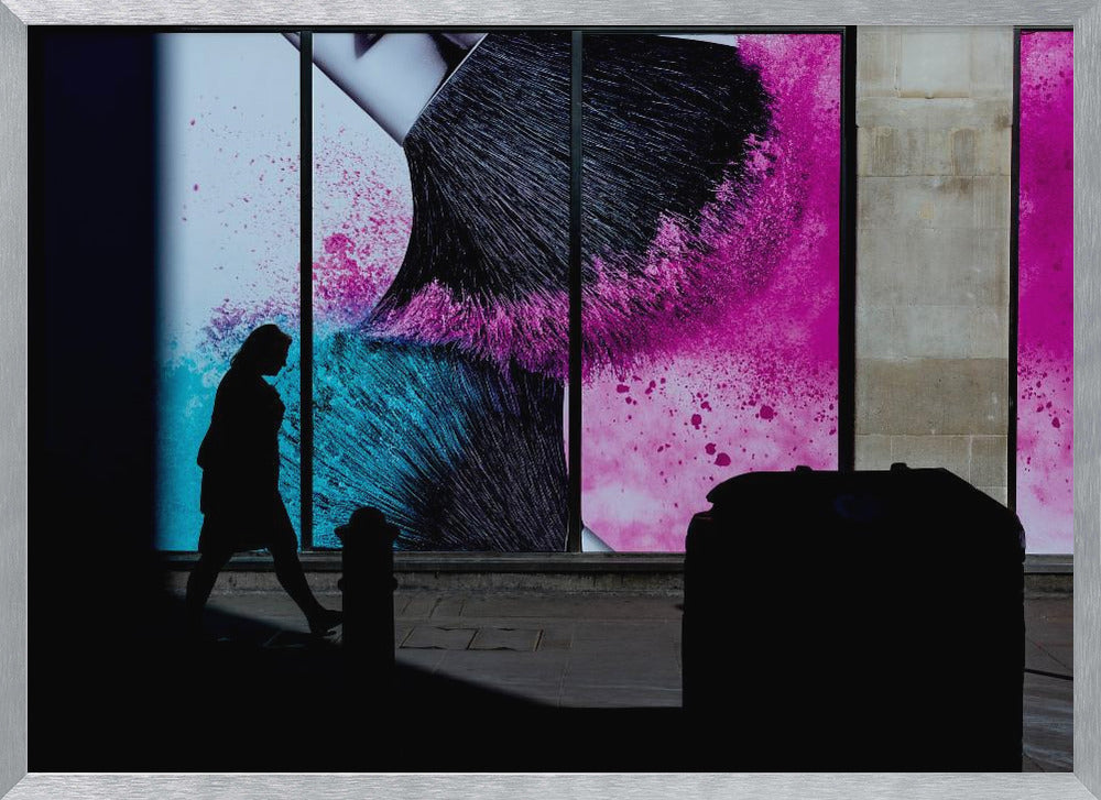 A framed photograph of a street scene where a person's silhouette is captured walking past a large, vibrant window advertisement. The ad features a close-up of a makeup brush with explosions of magenta and cyan powder. The foreground is cast in deep shadow, creating a dramatic contrast. Print