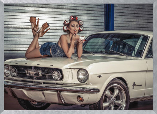 A woman in a retro pin-up style lies on the hood of a vintage white Ford Mustang in a garage, applying red lipstick with her hair set in soda can curlers. Poster