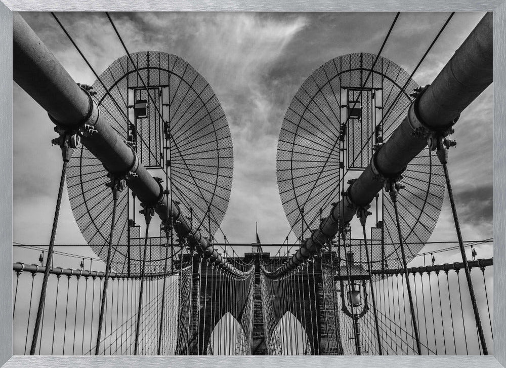 A symmetrical, low-angle black and white photograph of the Brooklyn Bridge's suspension cables and towers against a cloudy sky, framed in silver. Poster
