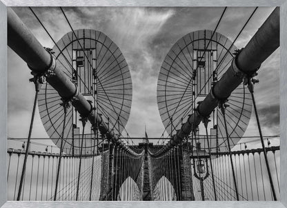 A symmetrical, low-angle black and white photograph of the Brooklyn Bridge's suspension cables and towers against a cloudy sky, framed in silver. Poster