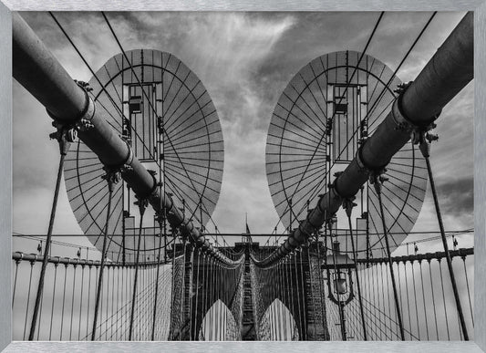 A symmetrical, low-angle black and white photograph of the Brooklyn Bridge's suspension cables and towers against a cloudy sky, framed in silver. Poster