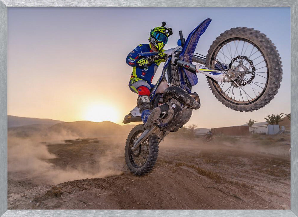 A motocross rider in blue and yellow gear is captured mid-air on a blue dirt bike, performing a jump against a dusty landscape with a setting sun in the background. The image is enclosed in a silver frame. Wall Art