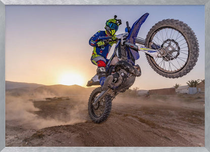A motocross rider in blue and yellow gear is captured mid-air on a blue dirt bike, performing a jump against a dusty landscape with a setting sun in the background. The image is enclosed in a silver frame. Wall Art