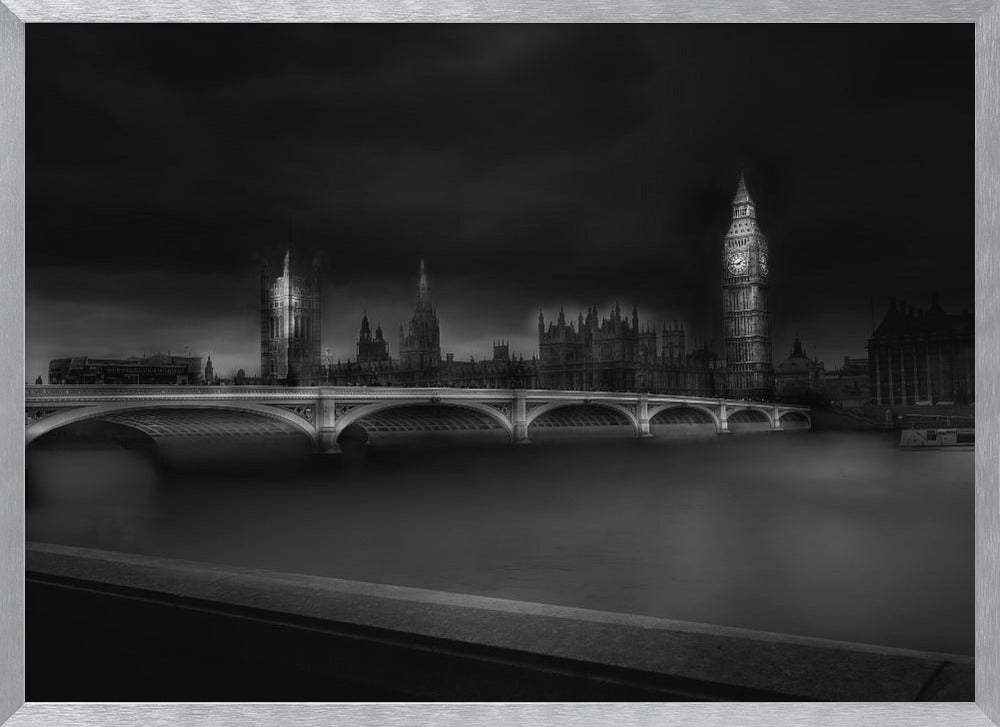 A moody, atmospheric black and white photograph of the Houses of Parliament and Big Ben in London, viewed from across the River Thames. The iconic buildings are softly illuminated against a dark, dramatic sky, with Westminster Bridge arching over the dark, smooth water in the foreground. The image is presented within a silver-colored frame. Artwork