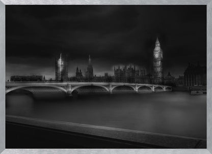 A moody, atmospheric black and white photograph of the Houses of Parliament and Big Ben in London, viewed from across the River Thames. The iconic buildings are softly illuminated against a dark, dramatic sky, with Westminster Bridge arching over the dark, smooth water in the foreground. The image is presented within a silver-colored frame. Artwork