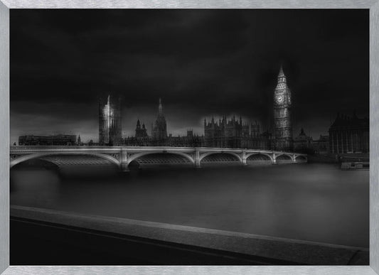 A moody, atmospheric black and white photograph of the Houses of Parliament and Big Ben in London, viewed from across the River Thames. The iconic buildings are softly illuminated against a dark, dramatic sky, with Westminster Bridge arching over the dark, smooth water in the foreground. The image is presented within a silver-colored frame. Artwork