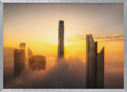 A framed photograph of a modern city skyline with skyscrapers emerging from a thick layer of clouds at sunrise, creating a dramatic and ethereal scene against a golden sky. Wall Art