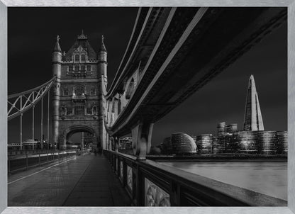 A dramatic, low-angle black and white photograph of London's Tower Bridge at night, taken from the pedestrian walkway. The image showcases the architectural details of the bridge and the illuminated modern city skyline, including The Shard, across the River Thames. Print