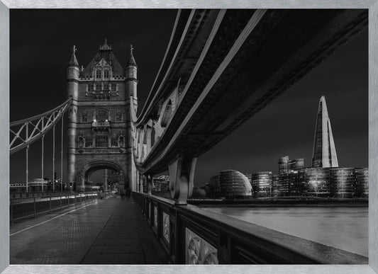 A dramatic, low-angle black and white photograph of London's Tower Bridge at night, taken from the pedestrian walkway. The image showcases the architectural details of the bridge and the illuminated modern city skyline, including The Shard, across the River Thames. Print