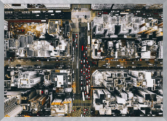 A framed, top-down aerial photo of a dense city grid. The monochromatic scene of white rooftops and dark streets is punctuated by the red and white cars creating lines of traffic below. Decor