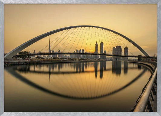A sweeping view of the Dubai Water Canal Bridge at sunset, its white arch and cables stretching over the calm water. The city skyline, including the Burj Khalifa, is silhouetted against the golden sky, with the entire scene creating a perfect reflection in the water below. Print