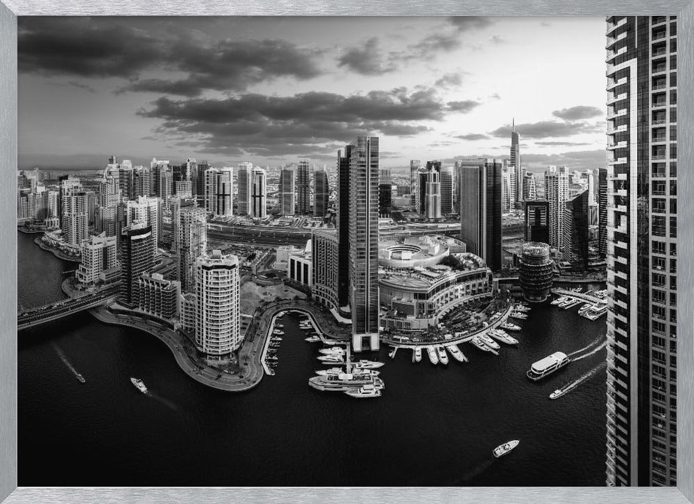 A framed, black and white panoramic photograph of the Dubai Marina taken from a high vantage point. The image captures the dense cityscape with numerous modern skyscrapers surrounding a winding waterway filled with docked yachts and moving boats. The sky is filled with dramatic, dark clouds, adding to the moody atmosphere of the urban landscape. Decor