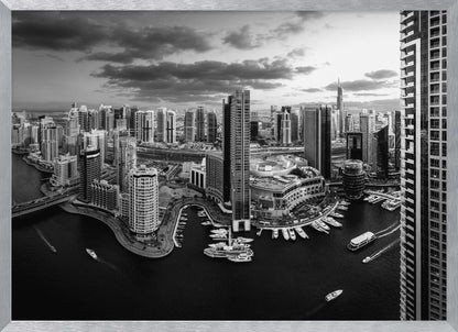A framed, black and white panoramic photograph of the Dubai Marina taken from a high vantage point. The image captures the dense cityscape with numerous modern skyscrapers surrounding a winding waterway filled with docked yachts and moving boats. The sky is filled with dramatic, dark clouds, adding to the moody atmosphere of the urban landscape. Decor