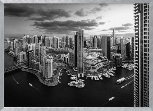 A framed, black and white panoramic photograph of the Dubai Marina taken from a high vantage point. The image captures the dense cityscape with numerous modern skyscrapers surrounding a winding waterway filled with docked yachts and moving boats. The sky is filled with dramatic, dark clouds, adding to the moody atmosphere of the urban landscape. Decor