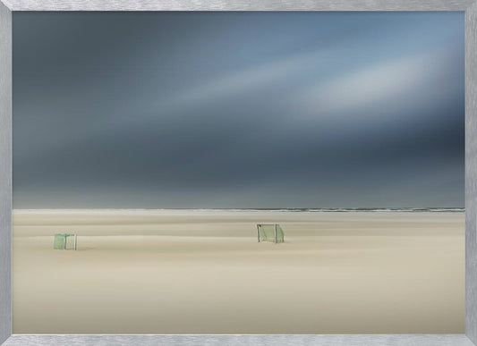 A minimalist long-exposure photograph of a vast, empty beach under a dark, moody sky. Two small, solitary soccer goals sit on the pale sand, with the ocean visible as a thin line in the distance. The artwork is presented in a silver frame. Artwork